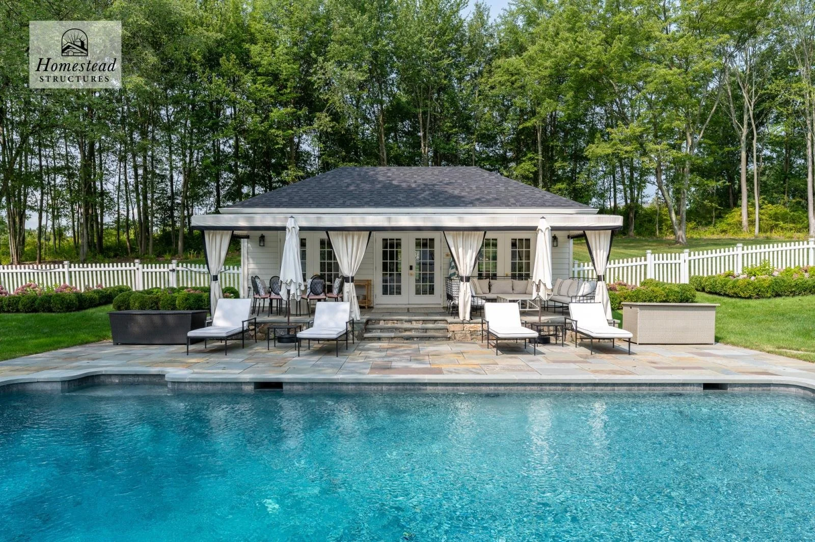 Backyard with swimming pool, outdoor patio furniture including lounge chairs, sofa, dining table with chairs, white curtains on a gazebo, surrounded by green grass, bushes, and trees, white picket fence, and a house in the background.