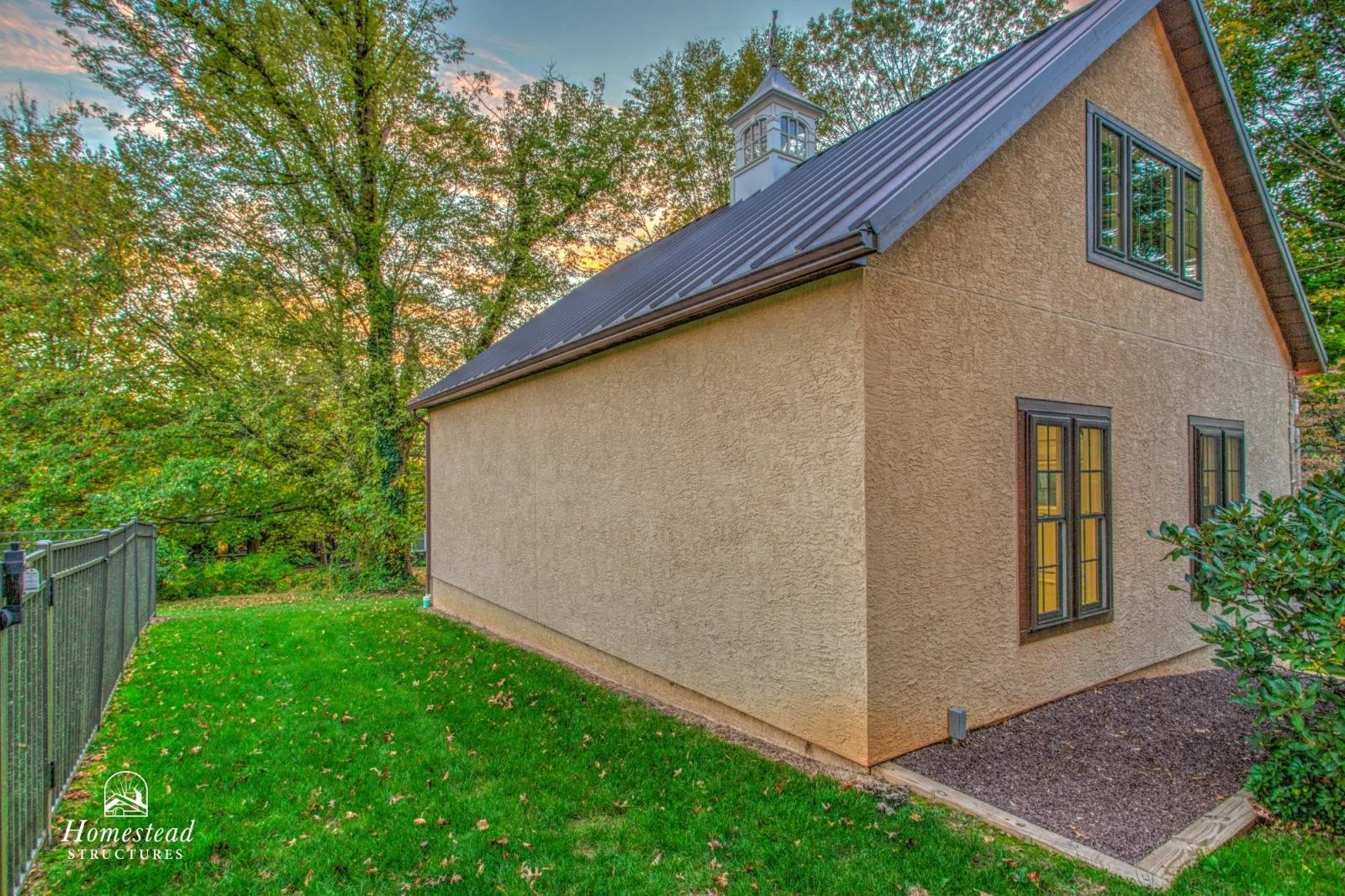 Side view of a beige stucco house with black window frames and a metal roof, surrounded by green grass, trees, and a dark metal fence, during sunset.