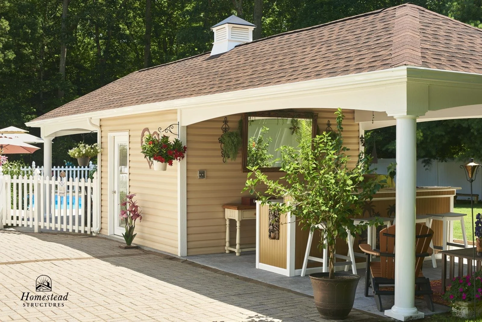 A small beige backyard structure with brown shingle roof, white trim, porch seating, hanging flower baskets, potted plants, and a white picket fence, surrounded by greenery.