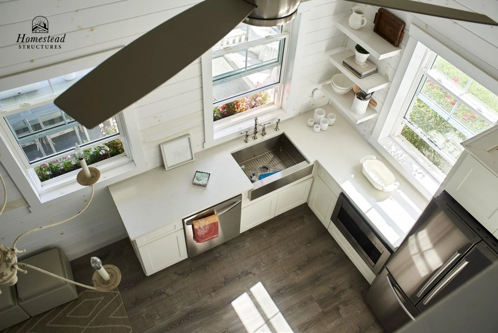 A white kitchen with three windows, a stainless steel sink, a microwave, a dishwasher, and a refrigerator, decorated with shelves and small kitchen items, illuminated by natural light.