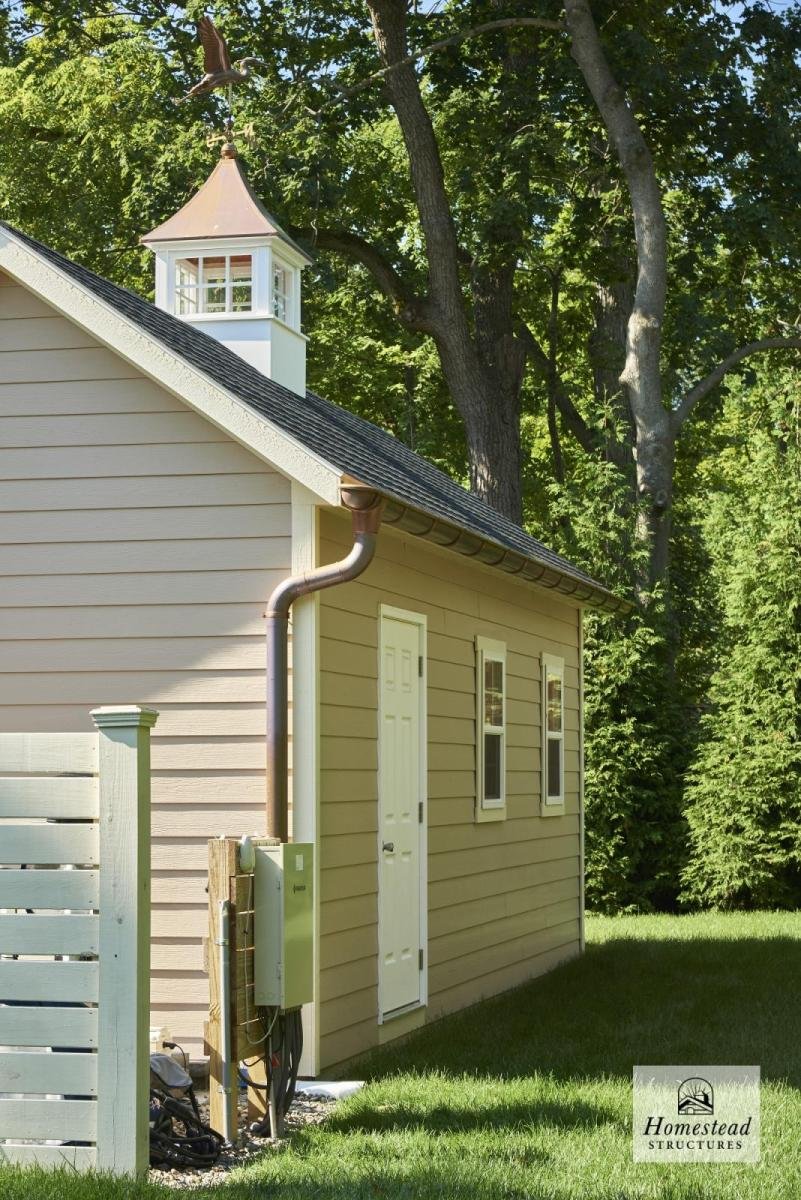 Side view of a beige house with a white door and two small windows, a black shingle roof, a small bell tower on top, surrounded by green trees and grass, with a wooden fence and utility box.