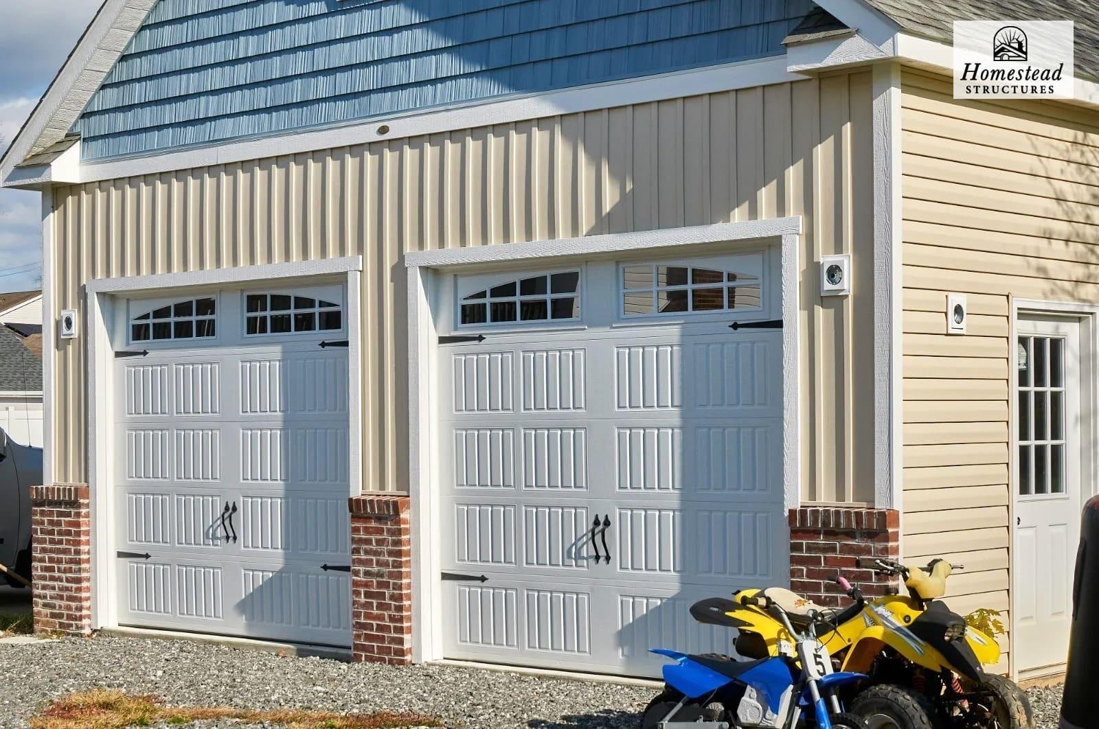 A two-car garage with two white doors, each featuring rectangular windows at the top. The garage has beige siding with brick accents at the base of the doors. Two motorcycles, one yellow and one blue, are parked in front of the garage. The structure 
