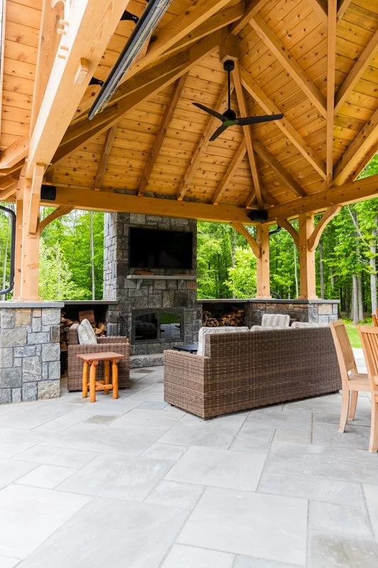 Outdoor pavilion with a wooden roof, ceiling fan, stone fireplace, flat-screen TV, and wicker furniture, surrounded by green trees.