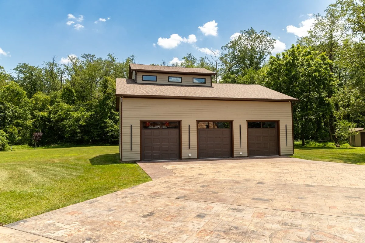 A large garage with three doors and a small upper window, situated in a grassy backyard surrounded by trees on a bright, sunny day.