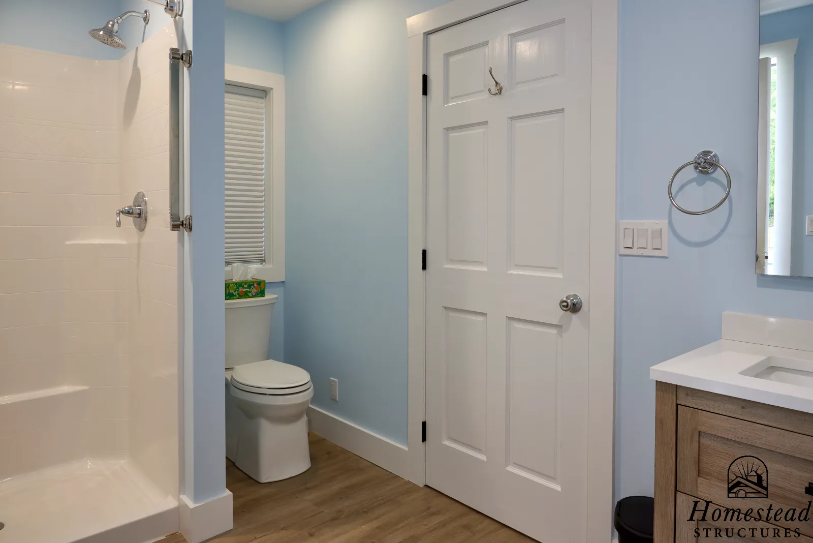 A bathroom with a shower stall on the left, a toilet in the center, and a wooden vanity with a sink on the right. The walls are painted light blue, and there is a white door with hooks on it. A mirror is partially visible on the right side.