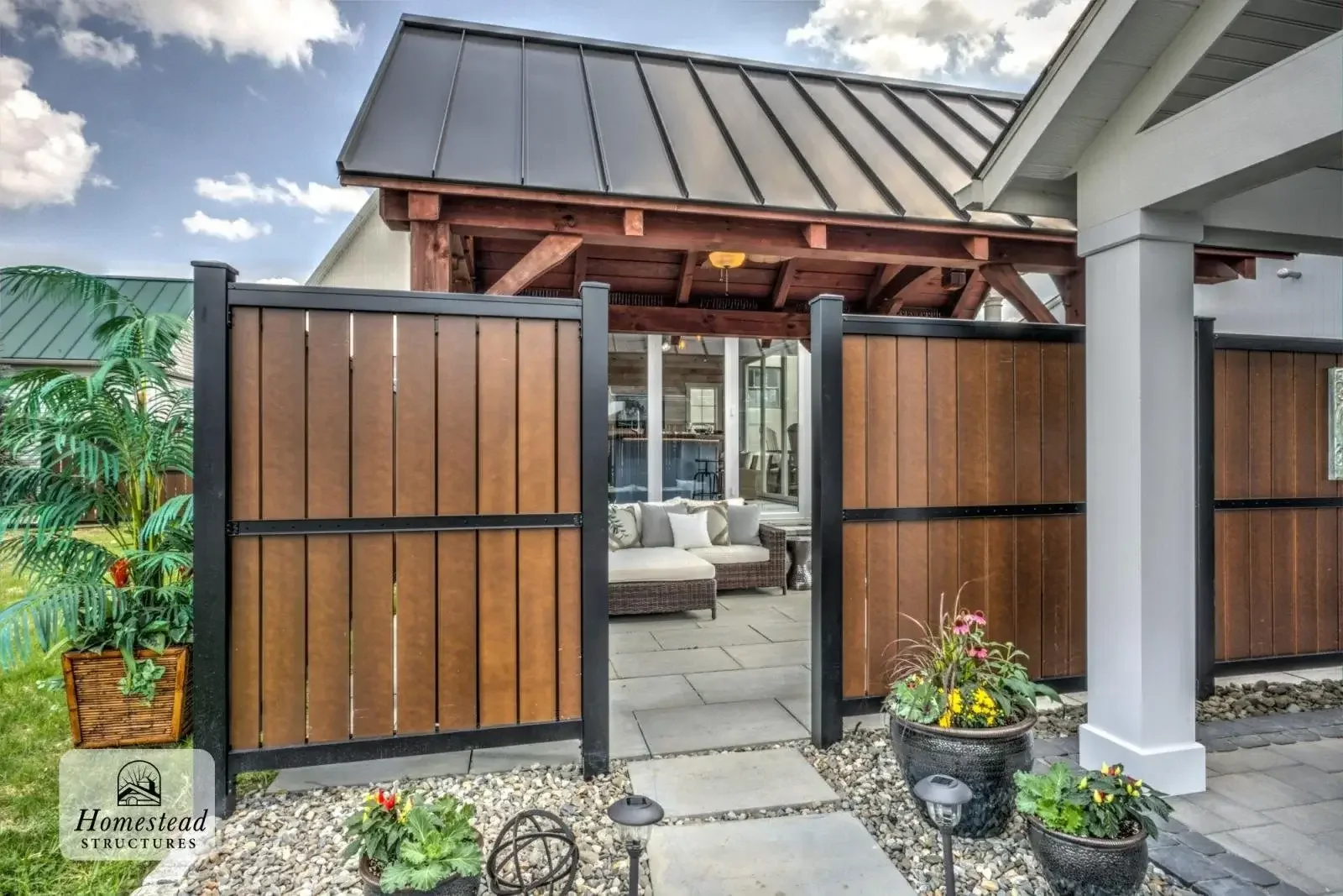 A backyard patio with a screened-off porch attached to a house, featuring outdoor furniture, a wicker armchair, a sectional sofa with cushions, potted plants, and a wooden pergola with a ceiling fan. The patio has stone flooring and steps leading to 