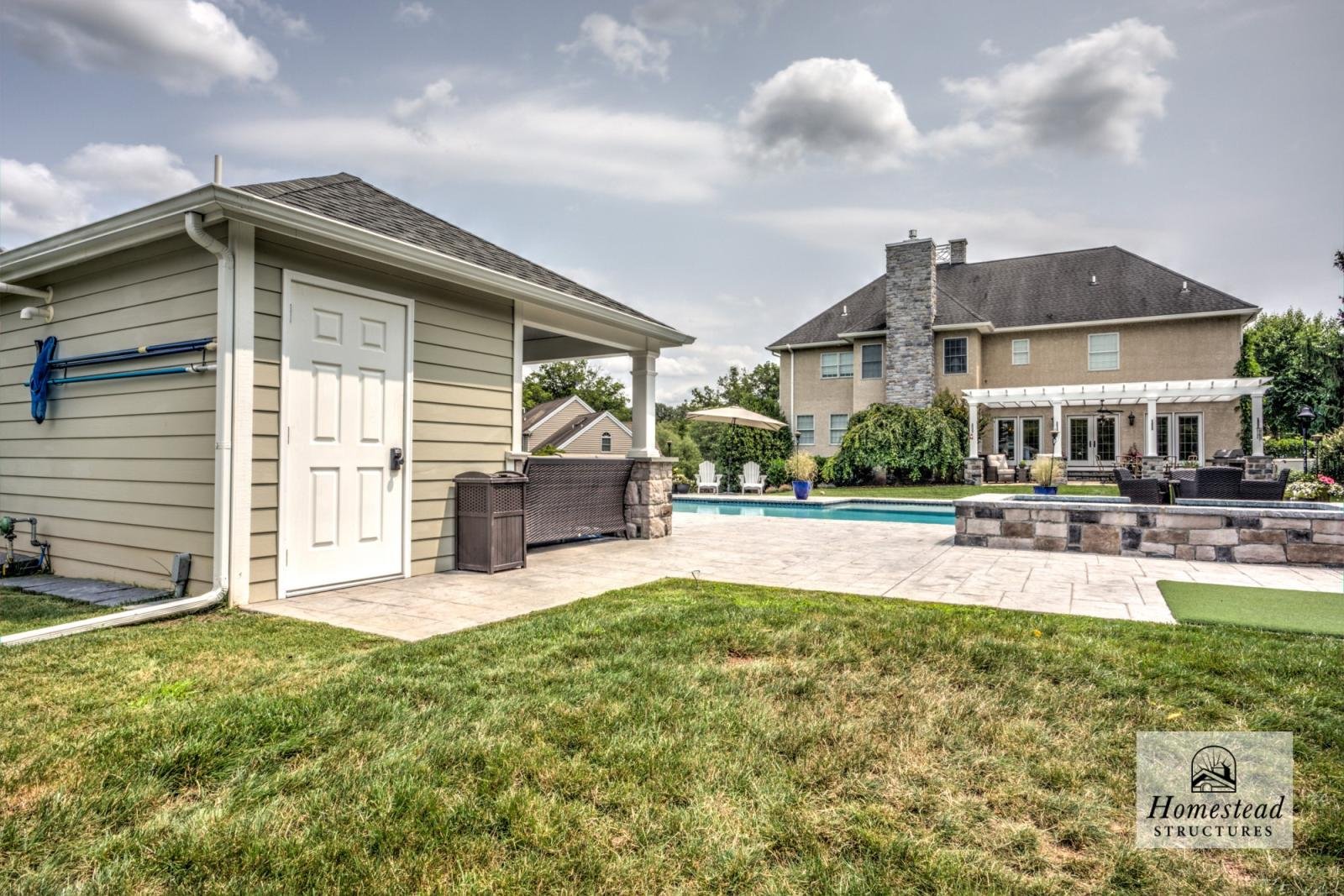 Backyard with a swimming pool, patio, outdoor furniture, a small beige building, and a large two-story house with a stone chimney, surrounded by greenery and trees.