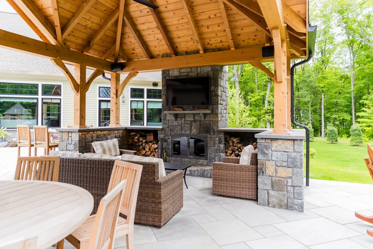 Outdoor covered patio with a stone fireplace, a mounted flat-screen TV, wicker chairs, a wooden dining table with chairs, and a view of lush green trees and grass.