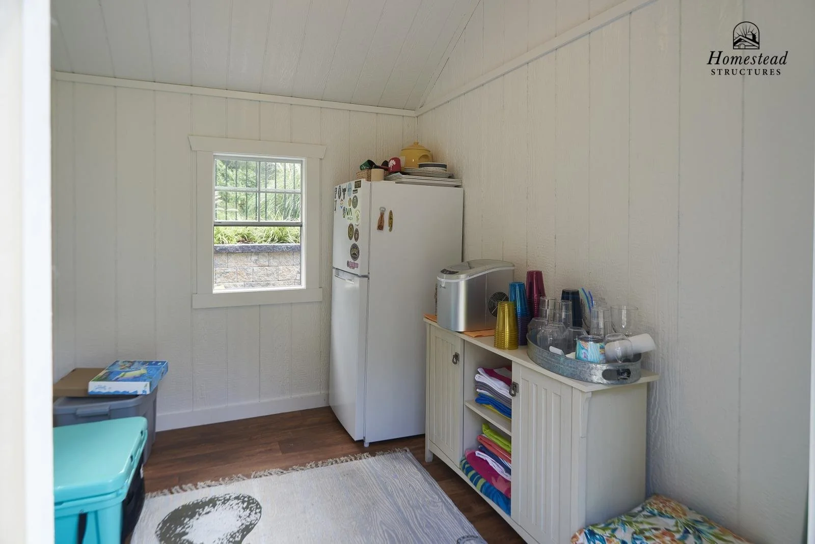Small room with a window, white walls, and a wooden floor. Contains a refrigerator with magnets, a white cabinet with colorful cups and glasses, and a tray of glasses and cups. There are stacked towels or cloths on the cabinet. The window shows green