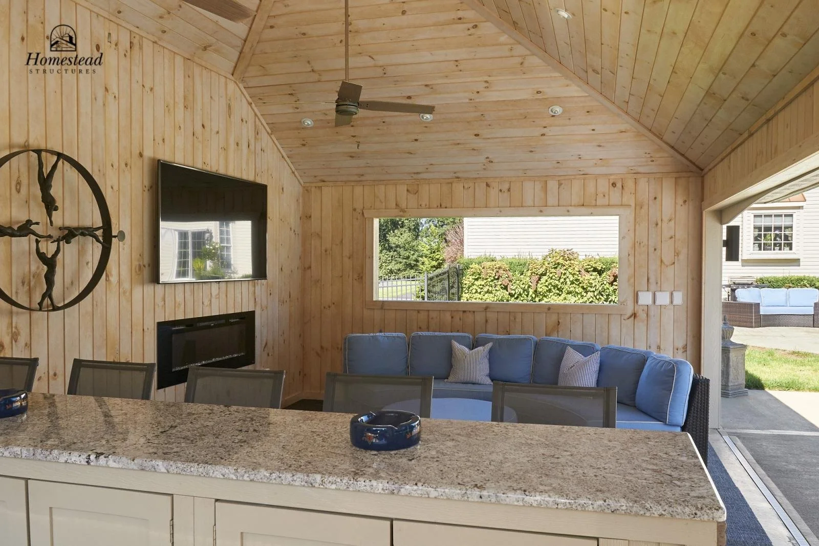Interior of a wooden room with a large window overlooking a garden, a blue sectional sofa, and a granite kitchen island in the foreground. There is a wall-mounted TV and a decorative wall sculpture.