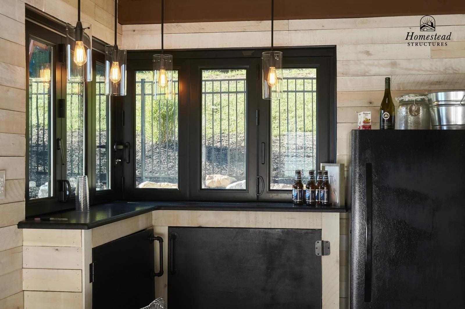 Interior of a rustic kitchen with black framed windows, wood panel walls, hanging pendant lights, a black countertop, and some bottles and containers on a black appliance.