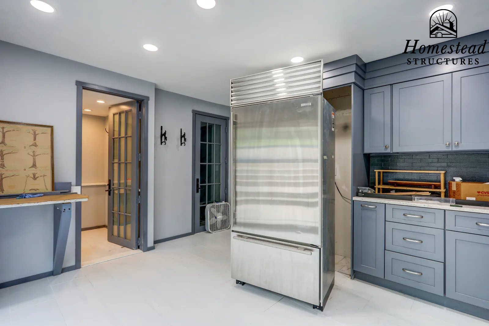 Kitchen with blue cabinets, stainless steel refrigerator, gray walls, French doors, and a small fan on the floor.