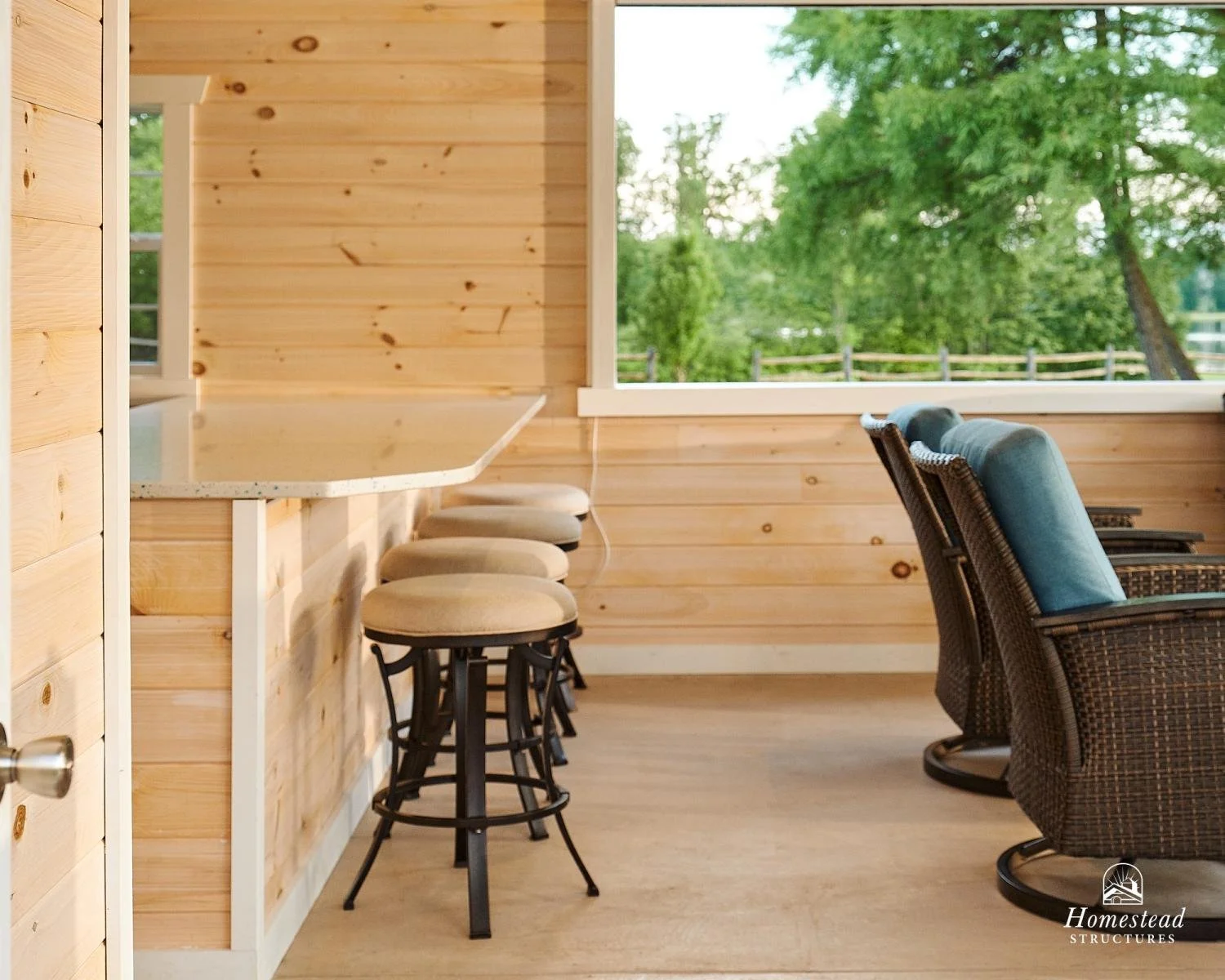 A cozy outdoor porch with wooden walls, a bar counter with stools, and wicker chairs with blue cushions overlooking lush green trees.