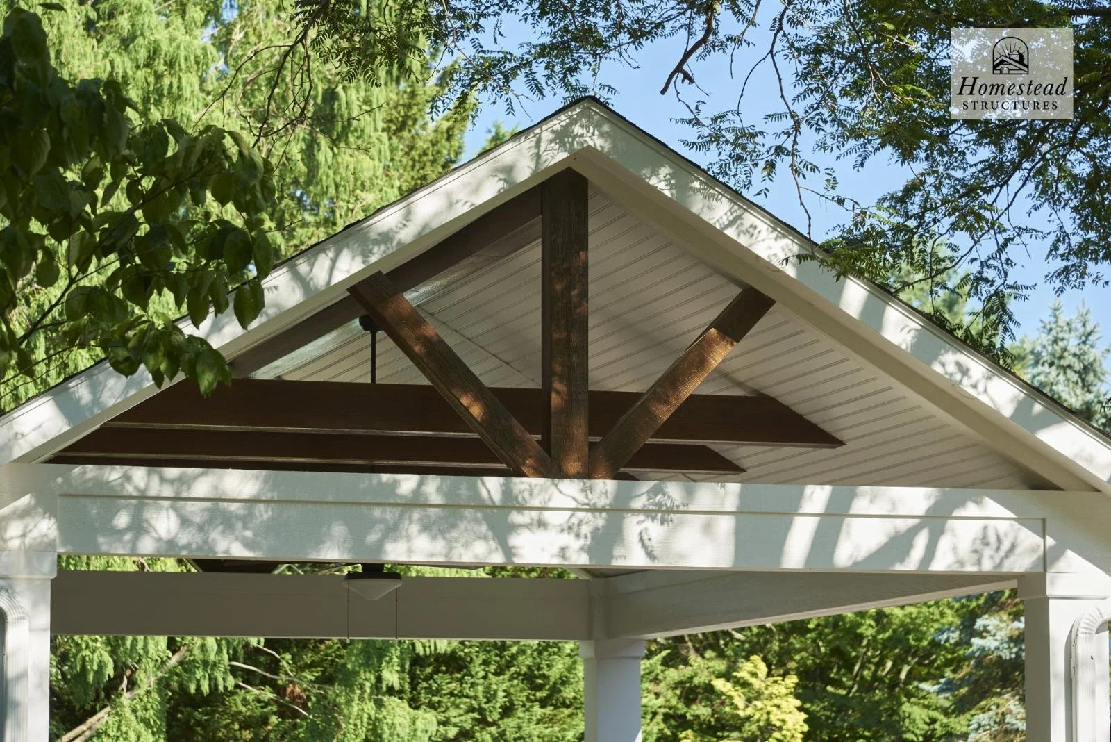 Close-up view of the underside of a white peaked roof with dark brown exposed beams, surrounded by green trees in a backyard.