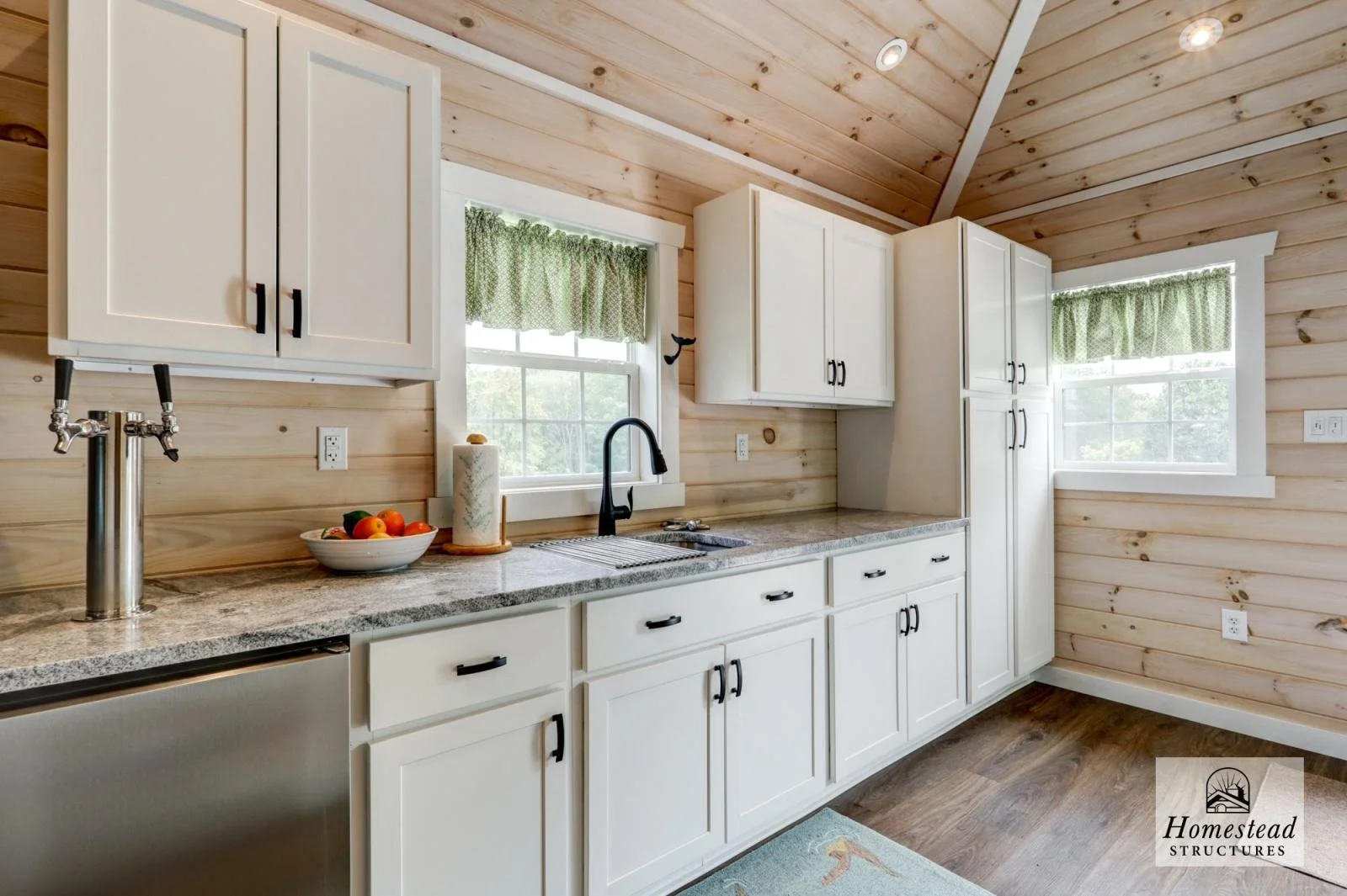 Kitchen with light wood wall paneling, white cabinets with black handles, granite countertop, black faucet, window with green curtains, and a bowl of fruit.