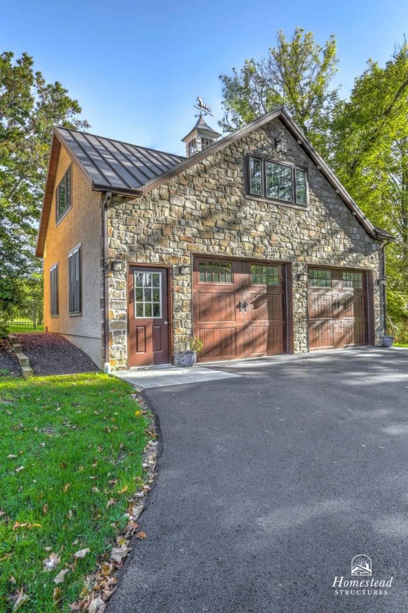 Stone and stucco garage with wooden doors, side door, and a small cupola with weather vane on roof, surrounded by trees and a paved driveway.