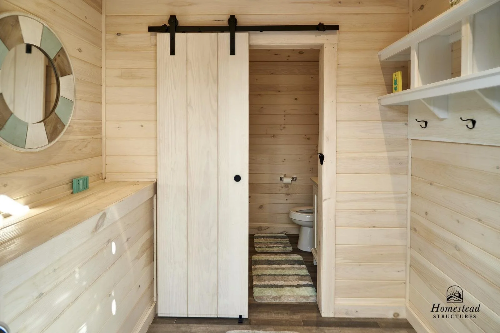 Interior of a small wooden bathroom with a sliding barn door, a toilet, a round mirror on the left wall, and shallow shelves on the right wall, with a colorful striped rug on the floor.