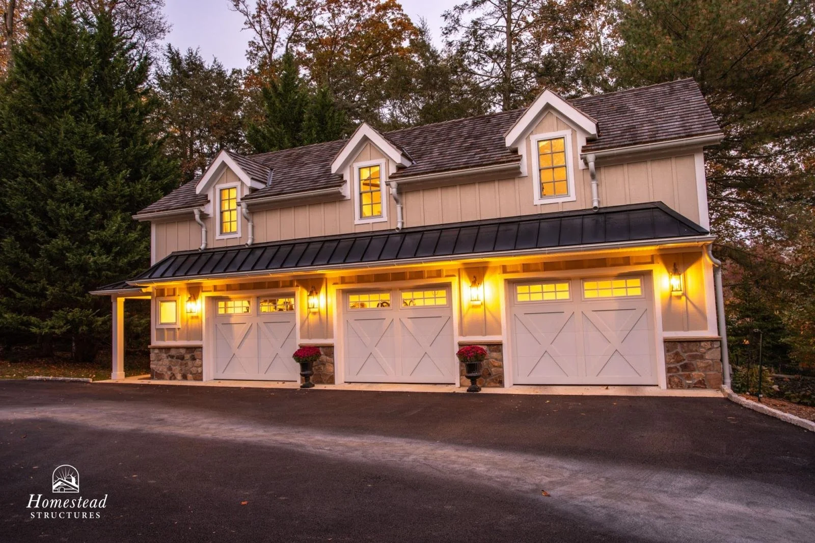A two-story garage with three white doors, stone accents, and yellow lighting, situated in a wooded area during dusk.