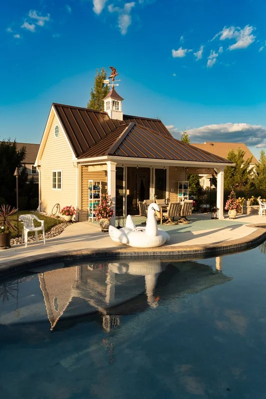 A backyard with a small house, a swimming pool, and pool floaties. There's outdoor furniture and potted plants, with a blue sky and clouds overhead.