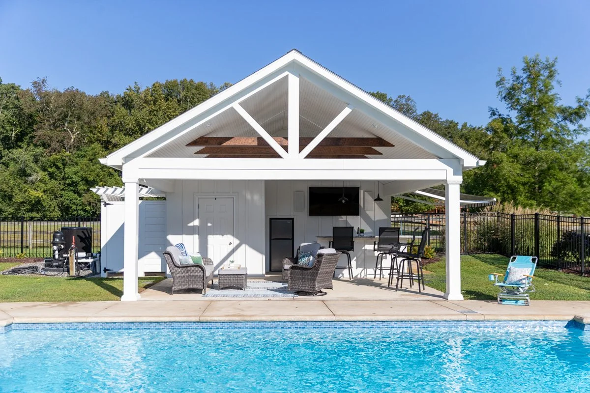 Outdoor pool area with a covered patio, lounge chairs, bar stools, and a small fridge, next to a swimming pool under a clear blue sky.
