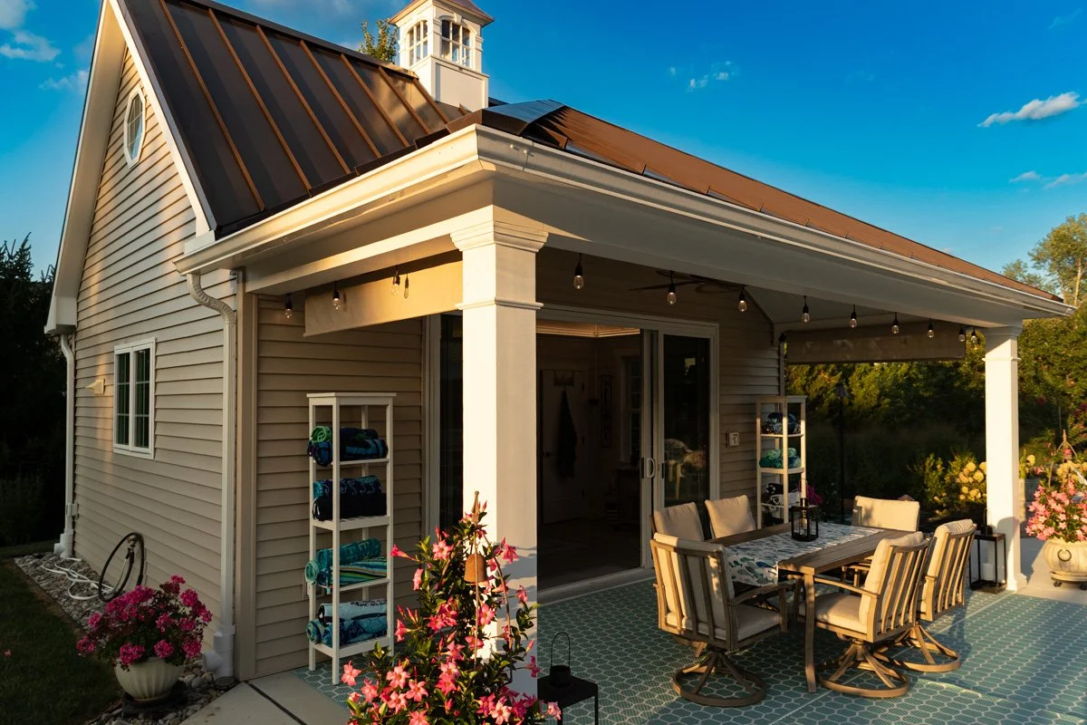 Backyard patio with a covered seating area, a dining table with chairs, flower pots, and a sliding glass door leading into a house with beige siding and a brown metal roof.