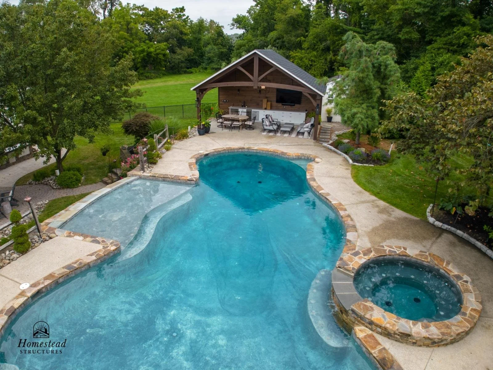 A backyard with a large, curvy swimming pool, a hot tub, and a covered outdoor living area with seating and a TV.