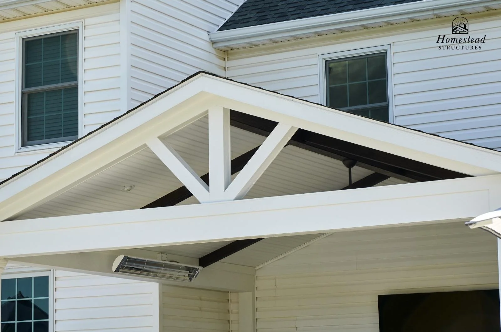 Close-up view of a white house exterior featuring siding, windows, and a covered porch with a white roof and a decorative truss, with a logo for Homestead Structures in the top right corner.