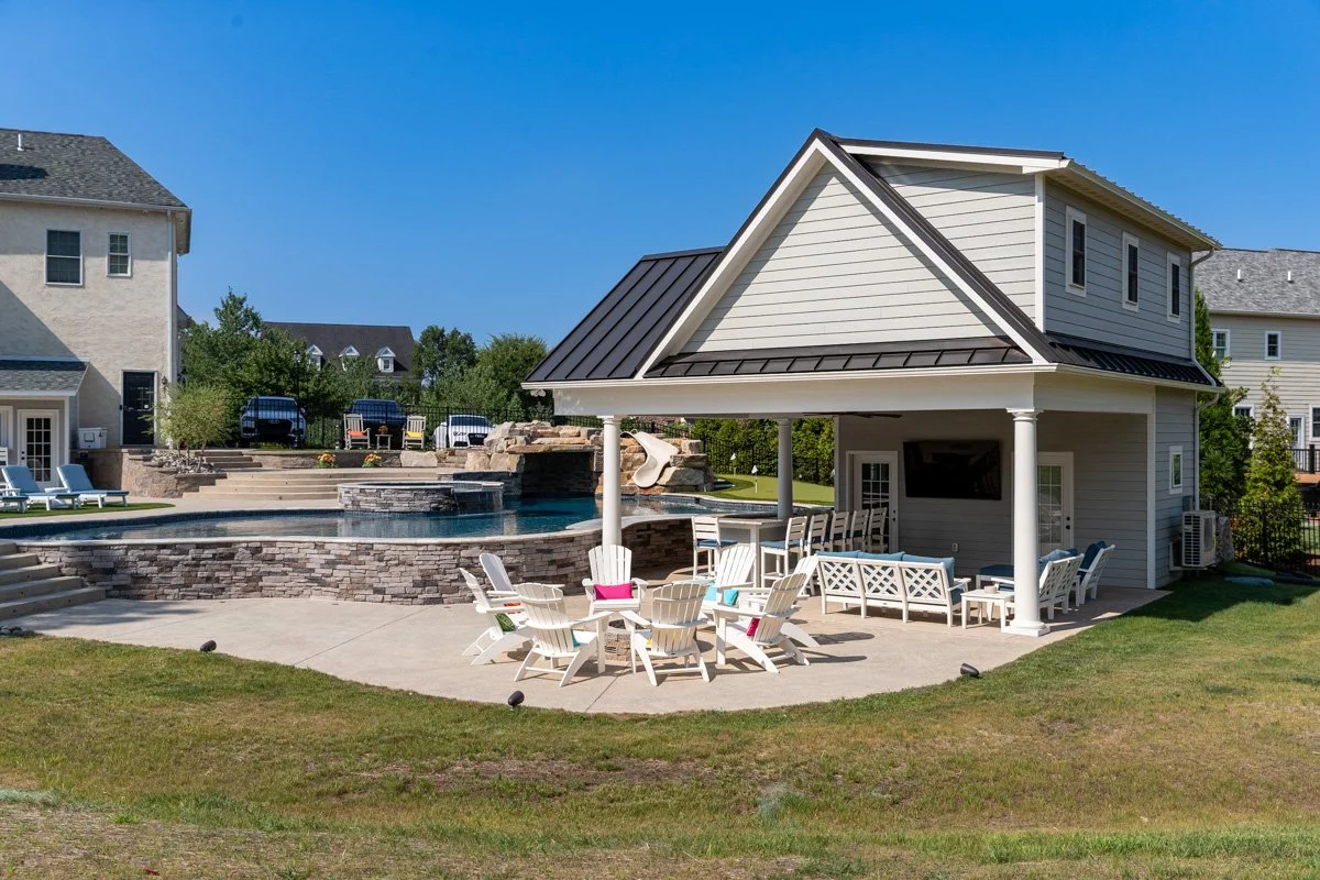 Backyard with a pool, waterfall feature, poolside lounge chairs, and a covered patio area with seating, surrounded by green grass and neighboring houses.