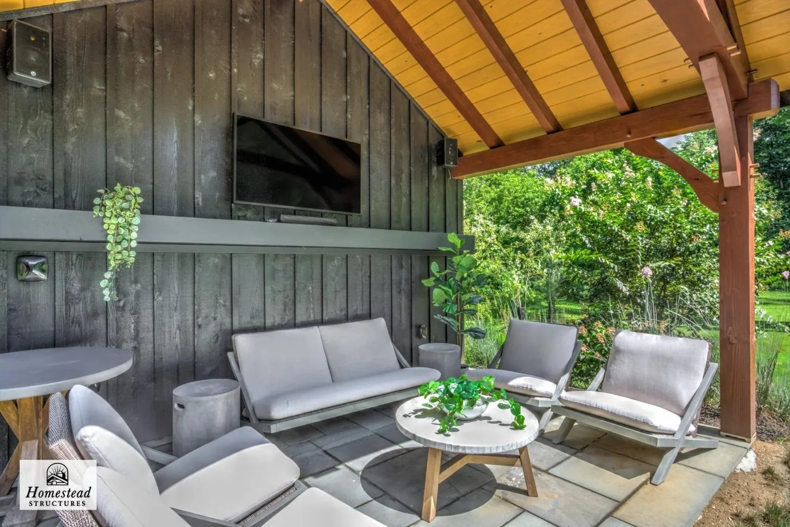 Covered outdoor patio with modern white furniture, a round coffee table with a plant, and a TV mounted on a dark wooden wall, surrounded by greenery.