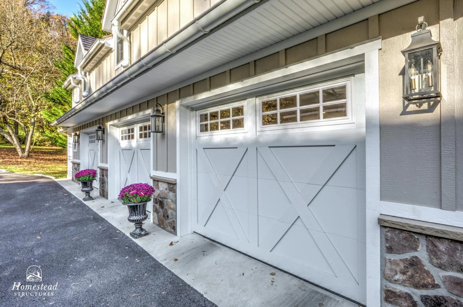View of a house garage with white carriage doors, stone accents, black lantern-style exterior lights, purple potted flowers, and a black asphalt driveway.