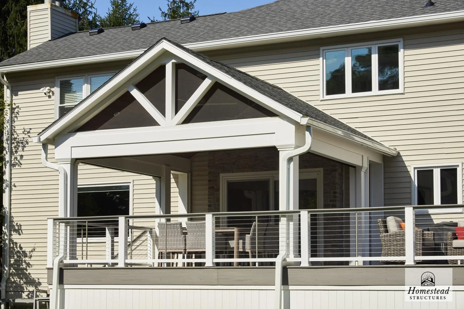 Newly constructed house with a covered porch, outdoor dining area, and seating on the deck, featuring beige siding, a gable roof, and white trim.