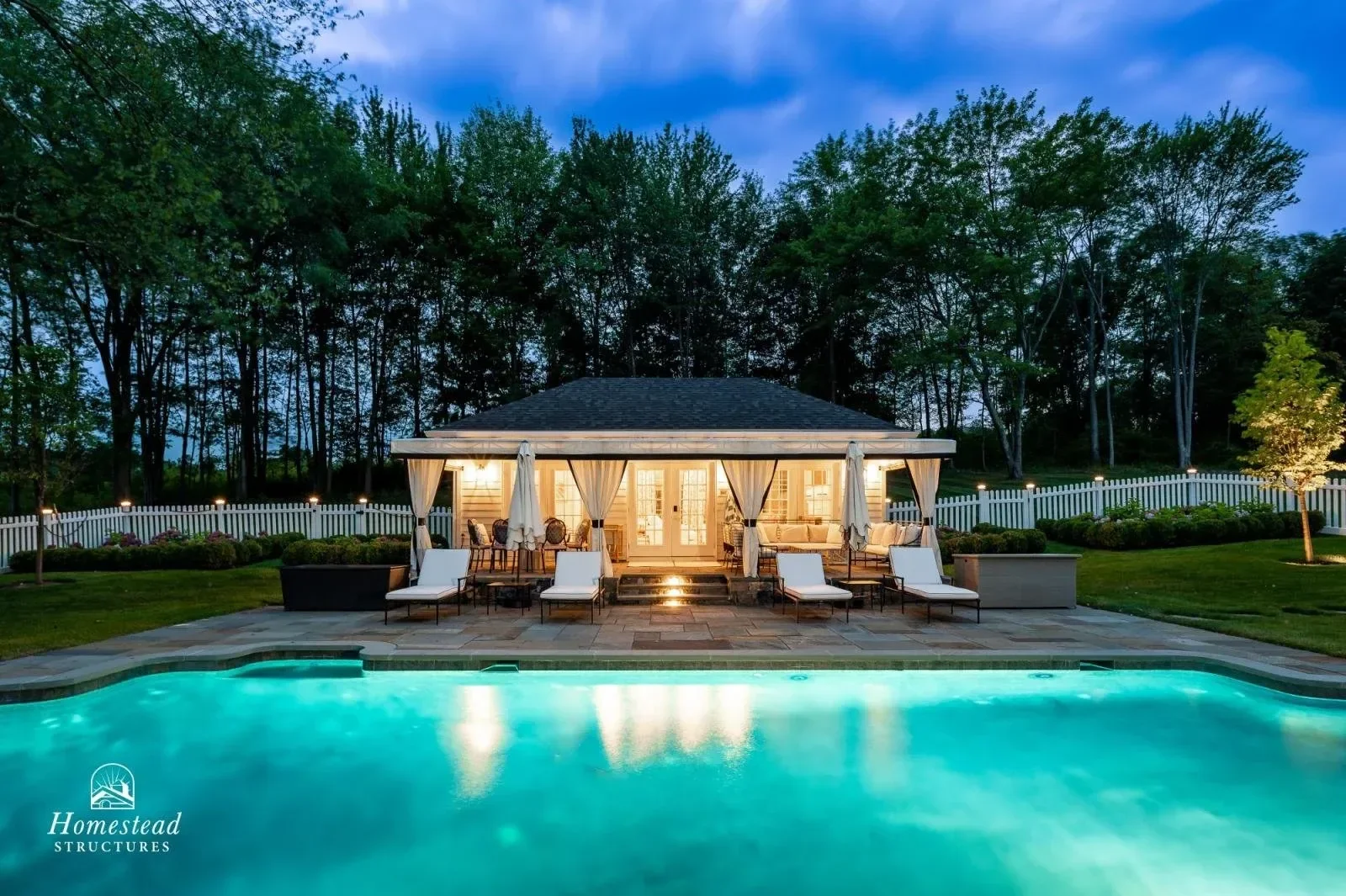 A backyard with a lit house, pool, lounge chairs, patio furniture, and a white fence, surrounded by trees, during evening.