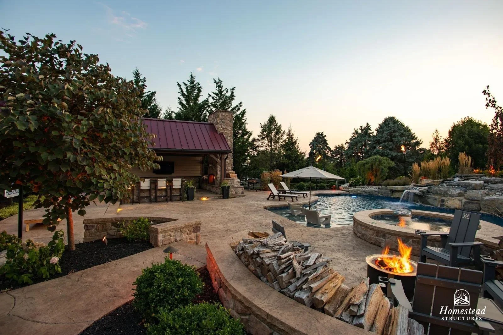 Luxury backyard with a swimming pool, fire pit, lounge chairs, a shaded umbrella, and a small outdoor bar with bar stools, surrounded by trees and landscaped garden in the evening.