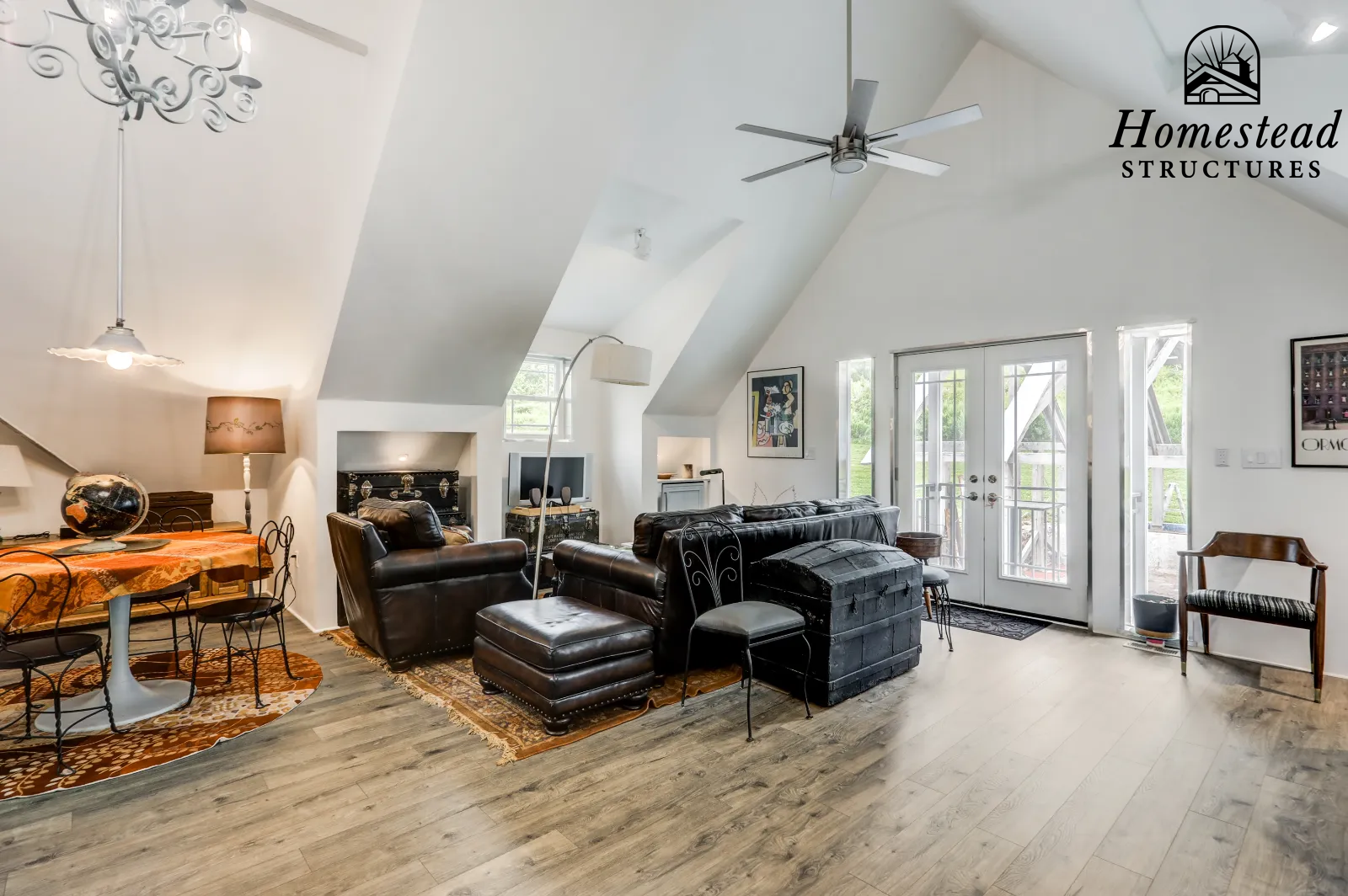 Living room with white sloped ceiling, ceiling fan, and natural light from glass doors, containing leather sofa and chairs, a dining table with chairs, art on the walls, and wooden flooring.