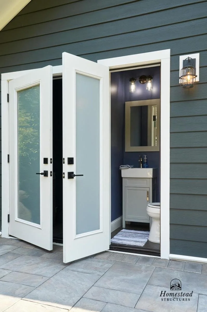 Outdoor view of a modern small bathroom with open bifold doors, blue interior walls, a small vanity with a mirror, a toilet, and exterior lighting fixtures.