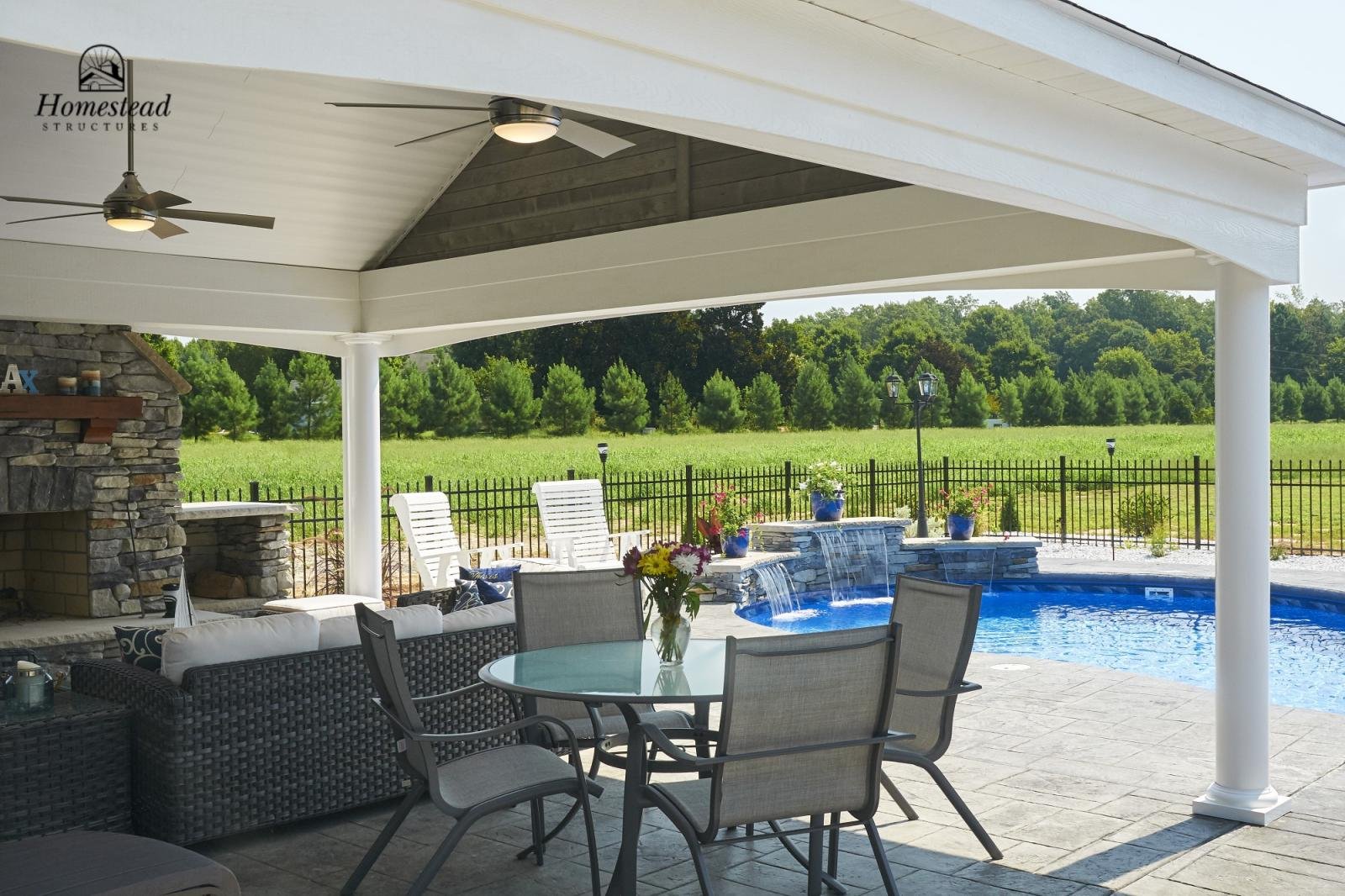 Covered patio with outdoor furniture, a water fountain, and a swimming pool, overlooking a green field with trees in the background.