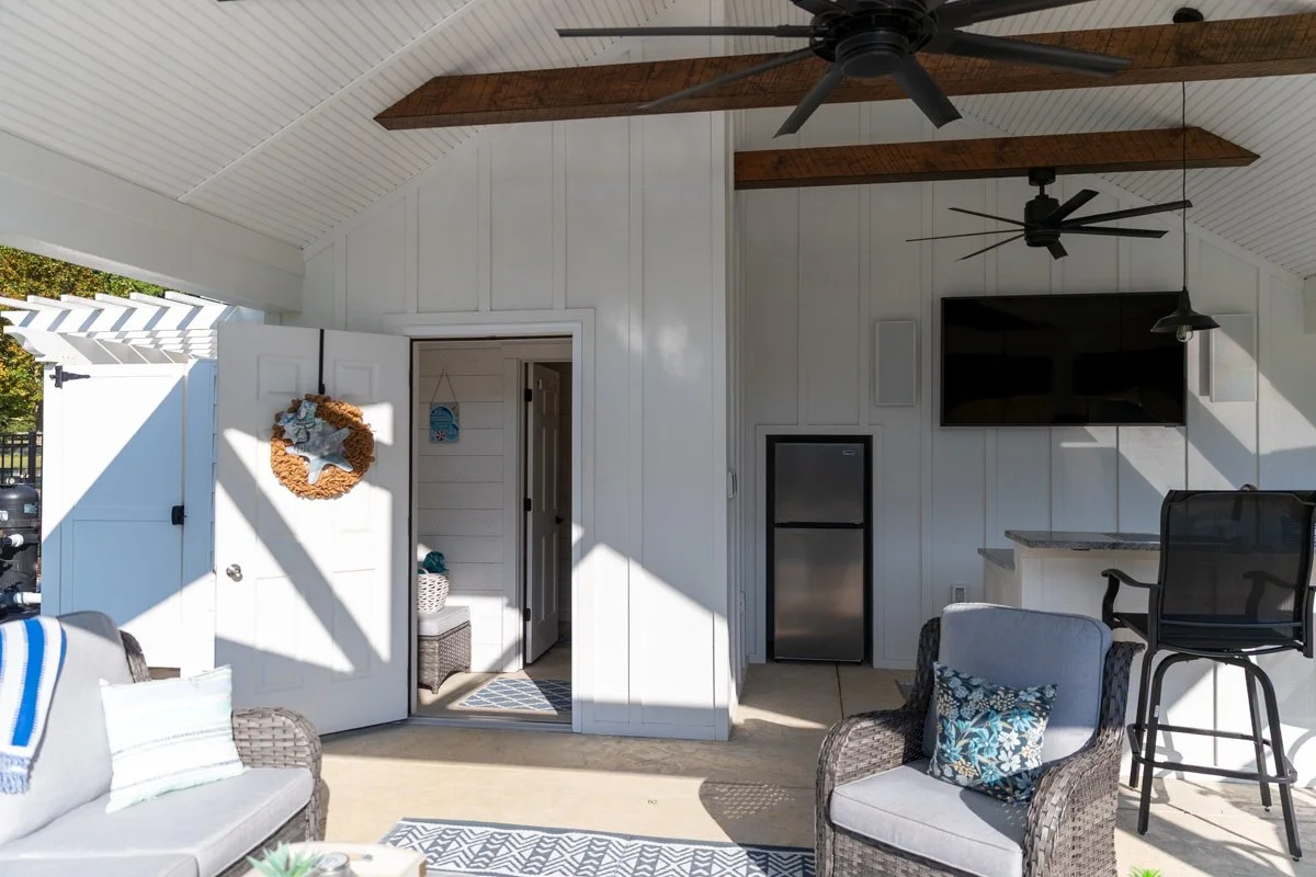 Covered outdoor patio area with wicker chairs, colorful pillows, ceiling fans, a wall-mounted TV, a small fridge, and various decor items, including a wreath on the door.