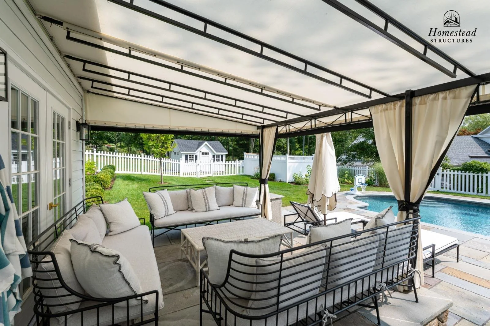 Covered outdoor patio area with beige furniture, including sofas and chairs with white cushions, overlooking a backyard with green lawn, white picket fence, a small shed, and a swimming pool.