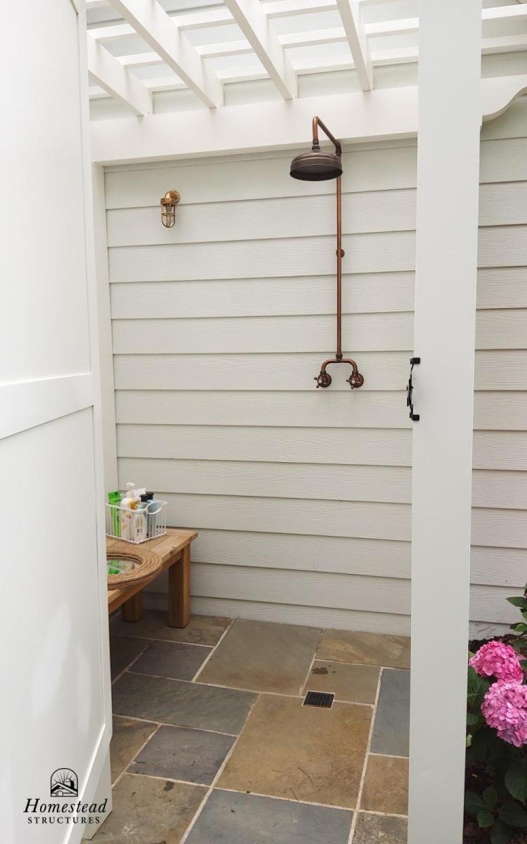 An outdoor shower with a black showerhead attached to a copper pipe on a white wooden wall, with a small wooden bench holding toiletries, and pink hydrangea flowers on the right.