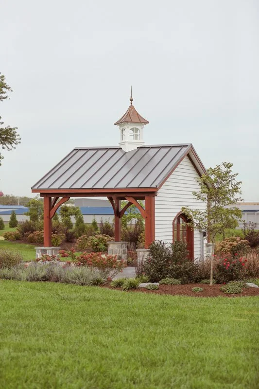 Small white chapel with a brown metal roof and a steeple, surrounded by greenery and colorful flowers.