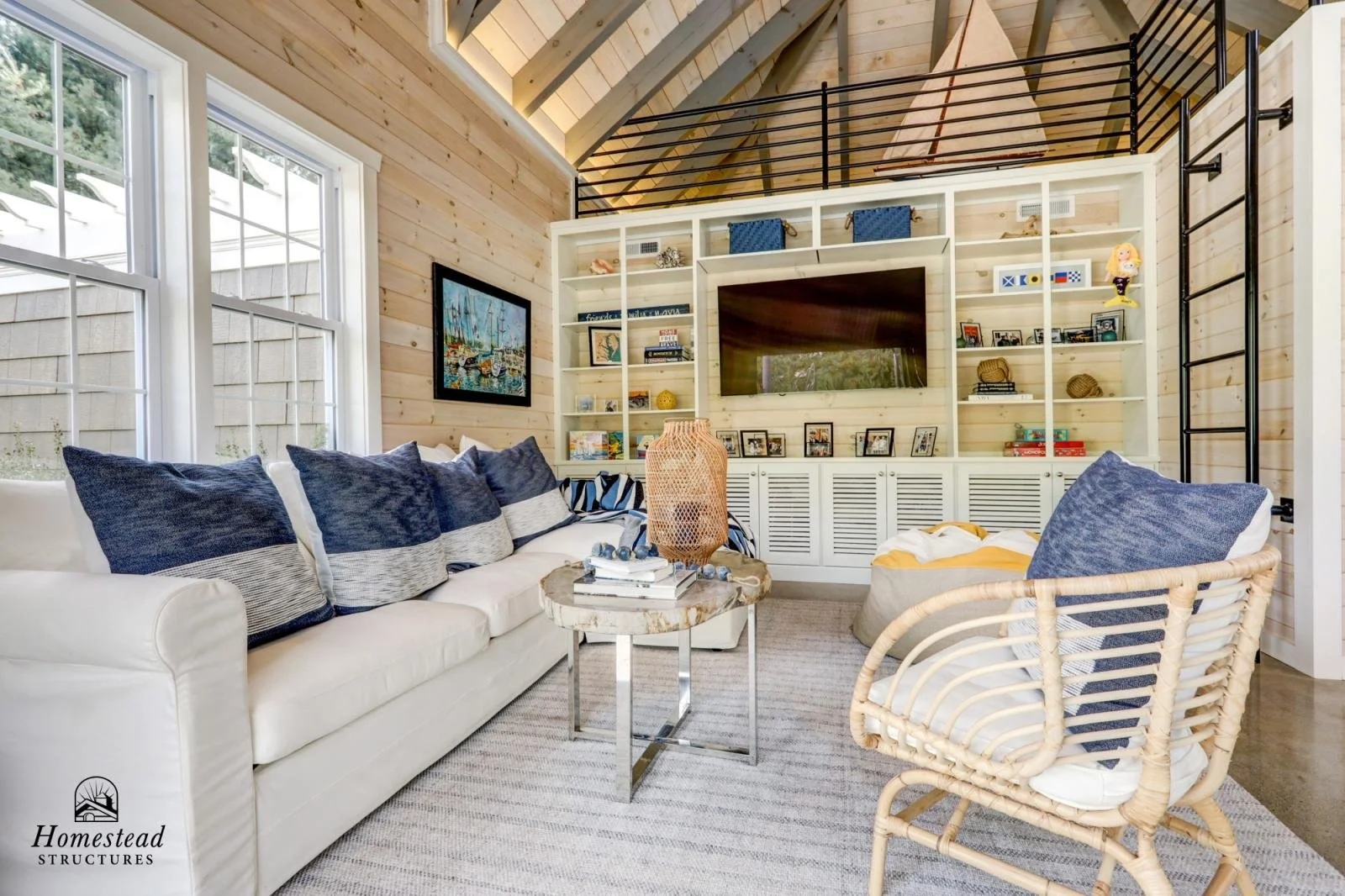 Cozy living room with a white sofa and a rattan armchair, decorated with blue and gray pillows. A small marble-top side table with a woven lamp and books is in front of the sofa. The room has light wood walls, a large window letting in natural light,