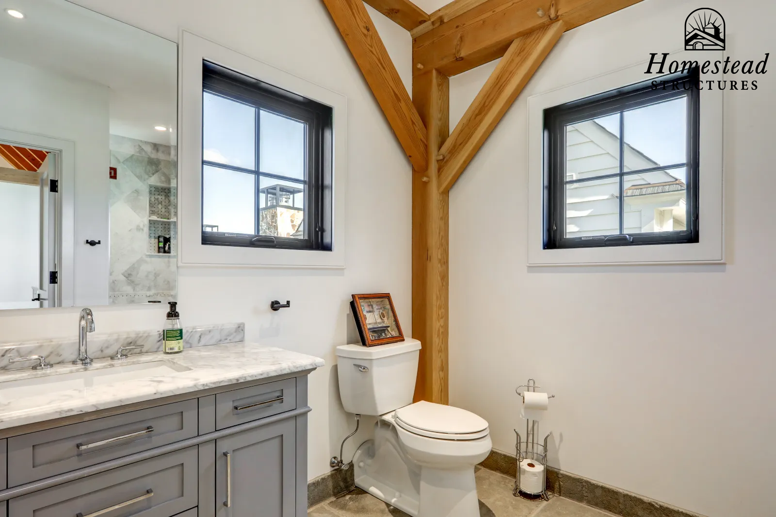 Bathroom with a gray vanity, marble countertop, two windows, and a white toilet. Wooden beams are visible in the ceiling. There is a framed picture on top of the toilet tank and a toilet paper holder with a spare roll on a stand. The walls are painte