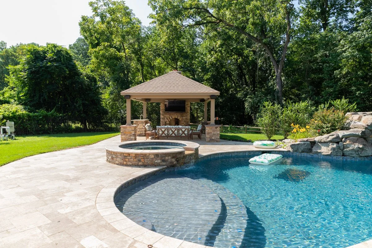 Backyard with swimming pool, hot tub, stone fireplace pavilion, surrounded by trees and greenery during daytime