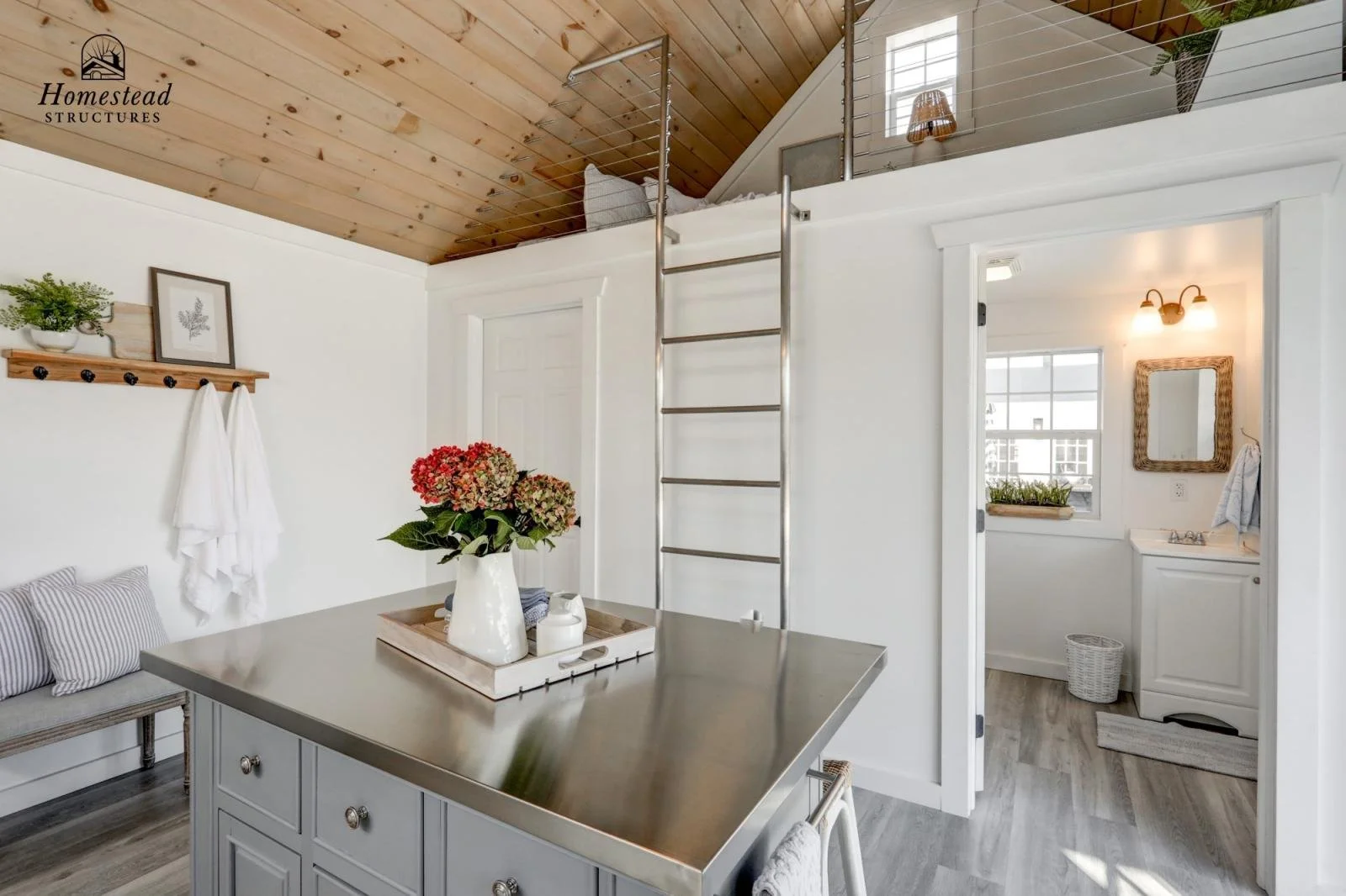 Interior of a cozy home with white walls, a kitchen island with a floral centerpiece, a wooden bench, and a loft bedroom accessible by a metal ladder. Includes a small bathroom with a mirror, window, and vanity.