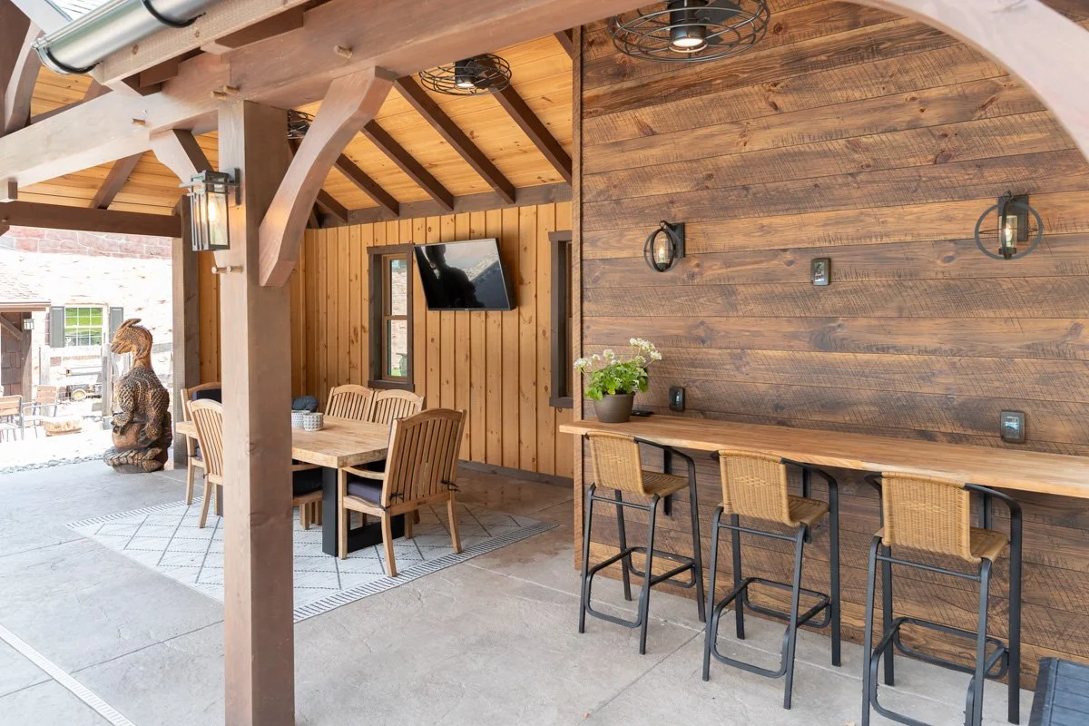 Indoor patio area with wooden ceiling and walls, featuring a dining table with chairs, a bar-height table with stools, wall-mounted lights, a mounted television, and decorative items including a plant and a wooden sculpture outside.
