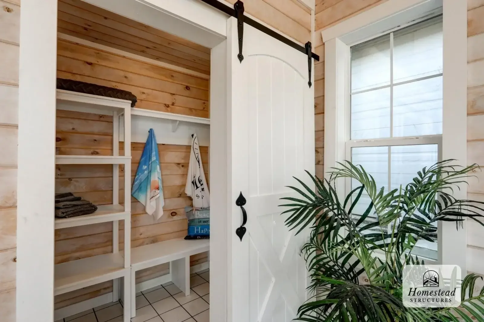 Interior of a small room with light wood paneled walls, white sliding door on a black metal track, a window with white trim, and a tall green potted plant. Inside, there are white shelves with towels and beach bags.