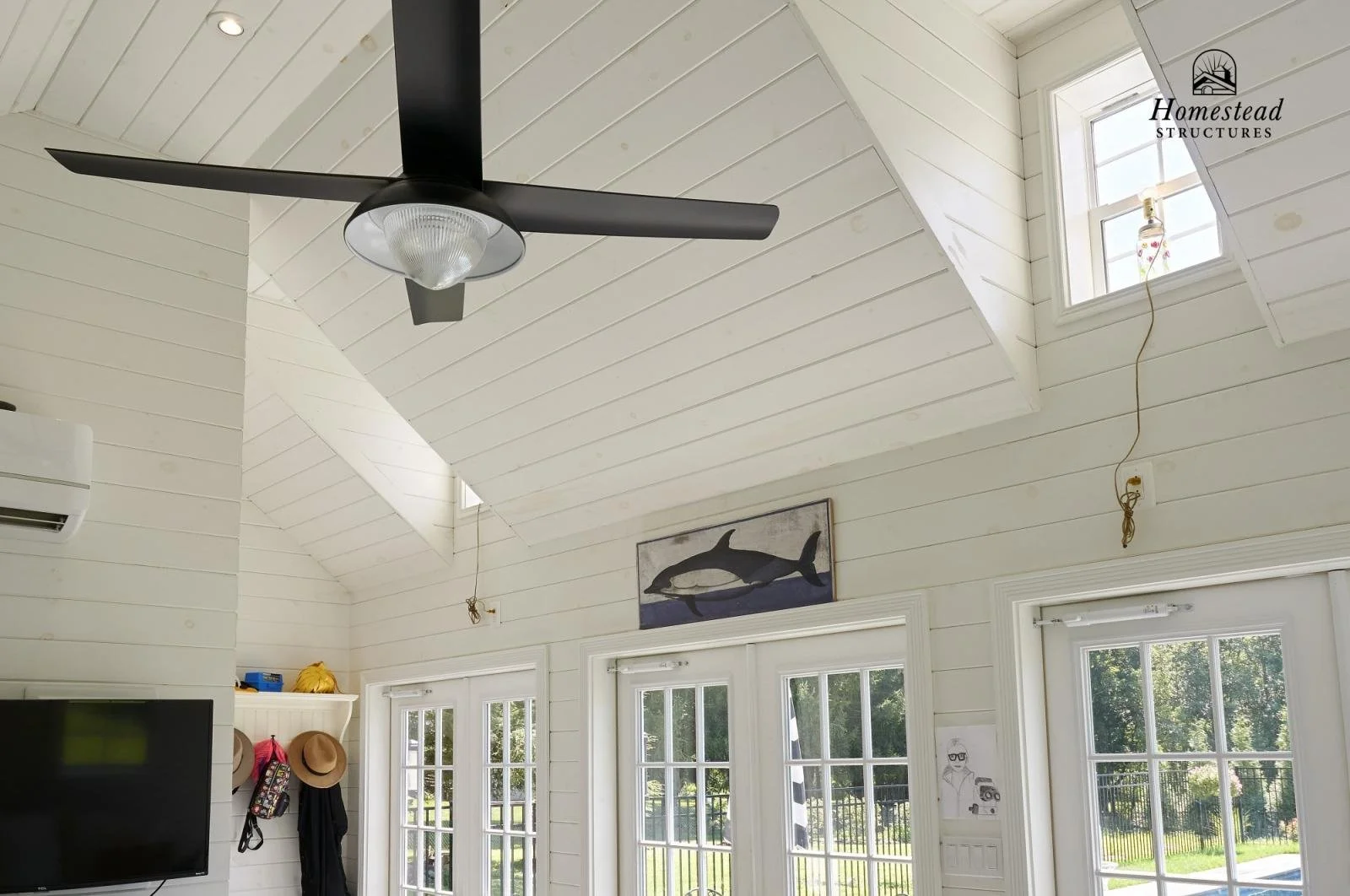 Interior of a room with white wooden walls and ceiling, three large windows showing greenery outside, a black ceiling fan, a fish painting on the wall, and a small drawing of a person with glasses and a camera near the door.