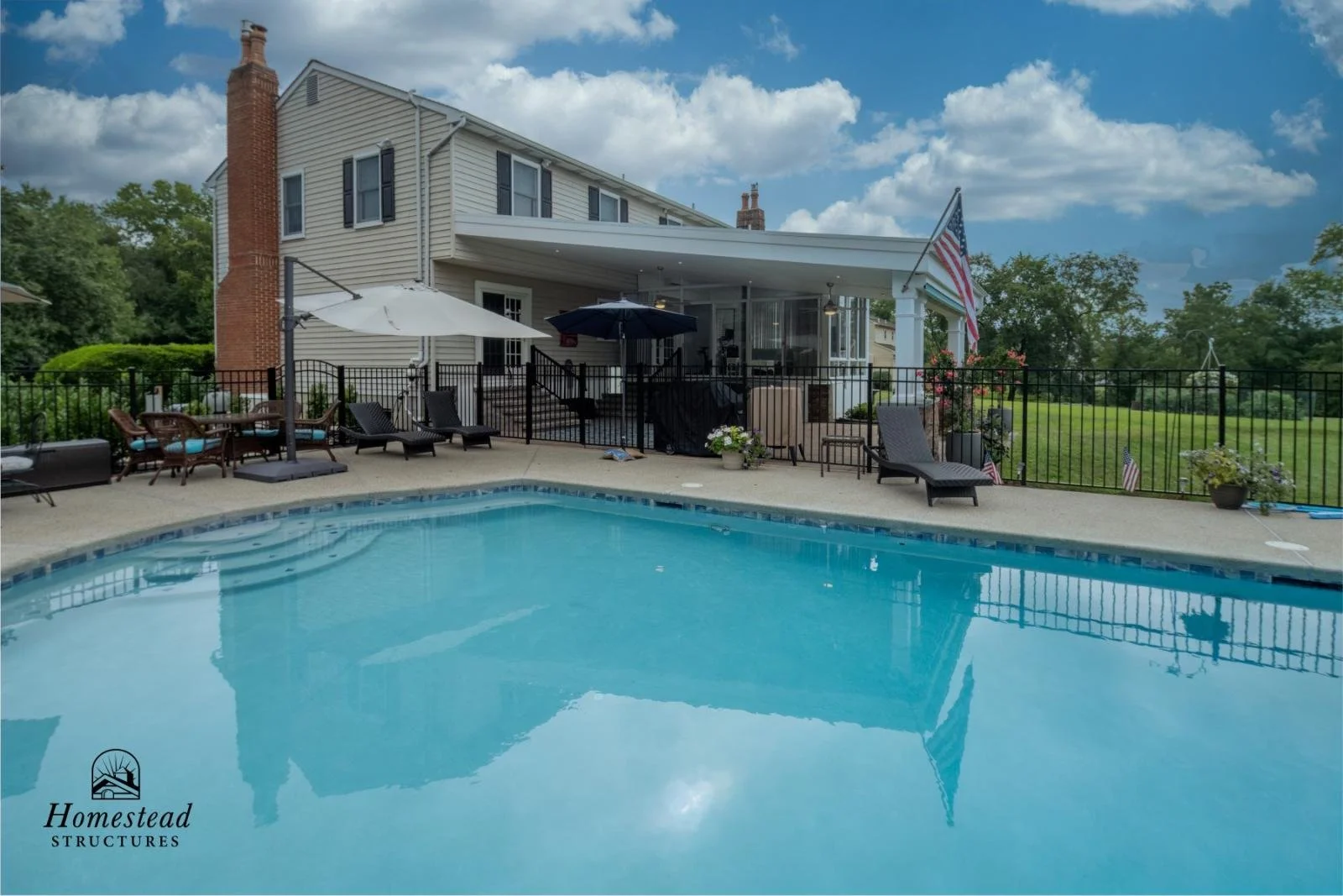 A backyard with a swimming pool, patio furniture, umbrellas, and a fenced yard, adjacent to a two-story house with beige siding, black shutters, and a brick chimney. There are American flags and potted plants around the area.