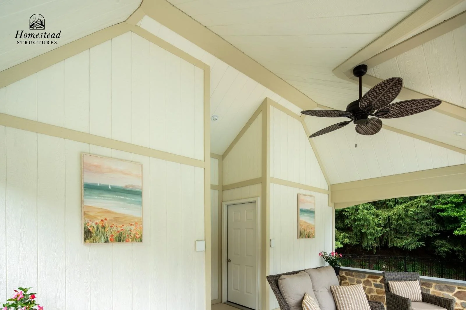Covered outdoor patio with white walls, a ceiling fan, a beige sofa with striped pillows, potted pink flowers, and landscape paintings, overlooking green trees.