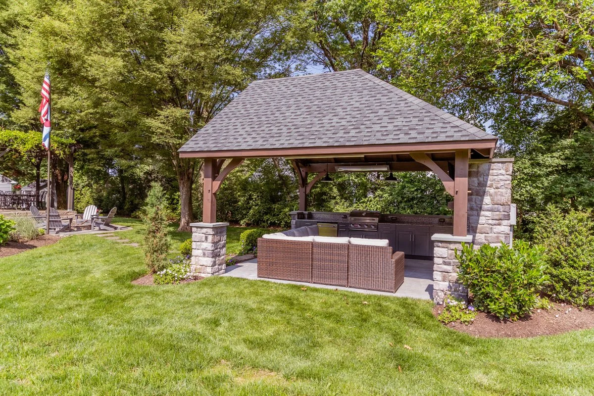 An outdoor patio with a small shelter featuring a gable roof, stone pillars, and built-in grill area with granite countertops. There is a wicker sectional sofa in front of the grill. The patio is surrounded by lush green grass, trees, and shrubs.